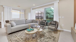 Ein modernes Wohnzimmer mit Ecksofa, Sesseln und einem Couchtisch. Große Fenster mit weißen Vorhängen, ein gemustertes Teppich und eine Deckenleuchte sind ebenfalls vorhanden.