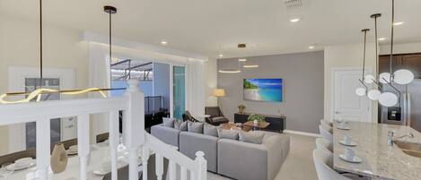 A modern kitchen with a central island, a dining area with a white railing, and a living room with a gray sofa and a TV.