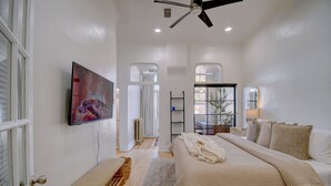 Une chambre avec un grand lit, un ventilateur de plafond, un téléviseur fixé au mur et une fenêtre avec des rideaux.