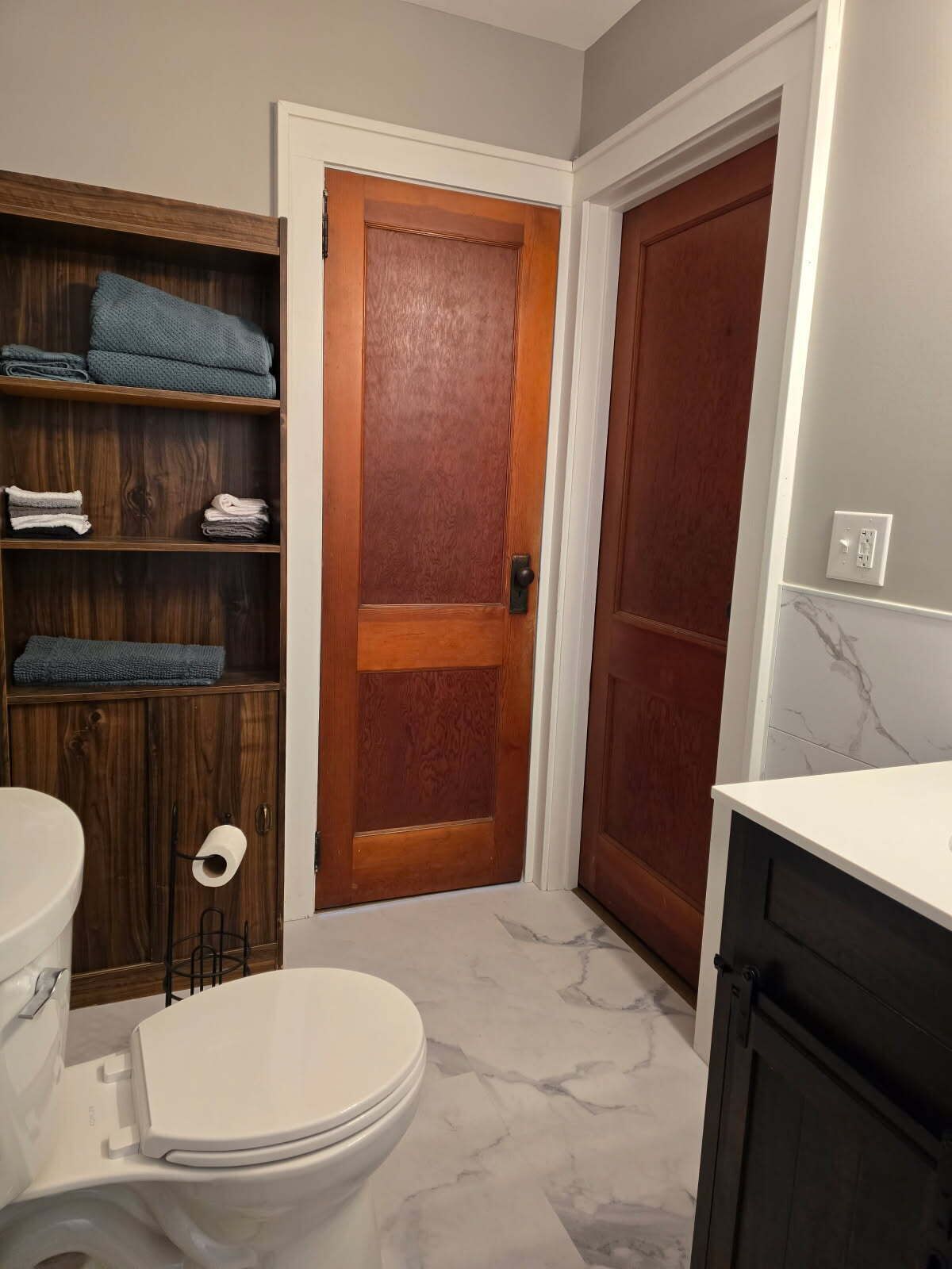 A bathroom with a toilet, a wooden shelf with folded towels, a marble floor, and a wooden door.