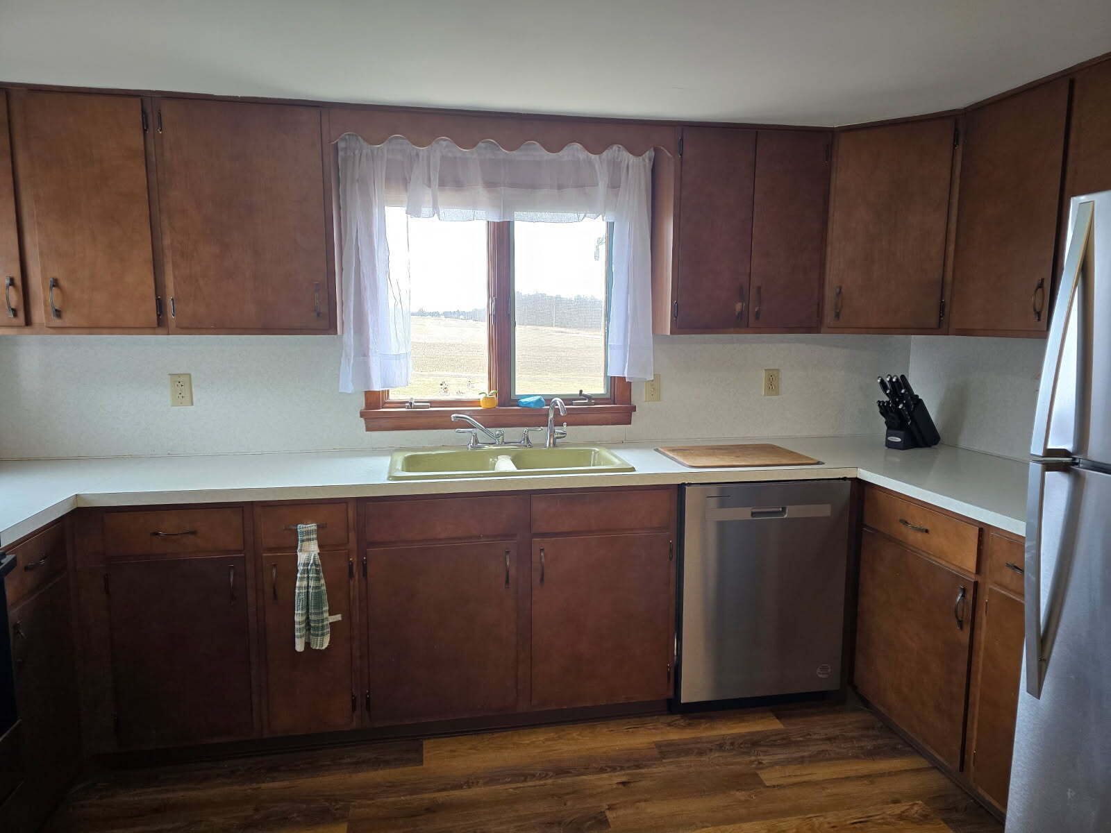 A kitchen with wooden cabinets, a stainless steel dishwasher, and a window with a white curtain.