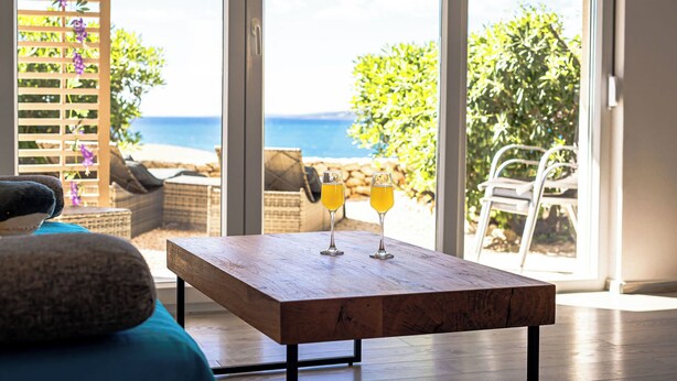 A wooden table with two glasses of orange juice, a wicker chair, and a view of the sea through large windows. - Quiet 79m2 apartment by the sea with garden and communal pool (Povljana)