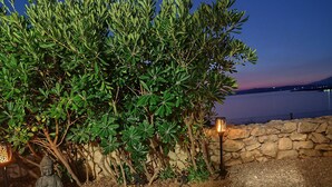 A serene outdoor seating area with a Buddha statue, a stone wall, and a view of the water at dusk. - Quiet 79m2 apartment by the sea with garden and communal pool (Povljana)