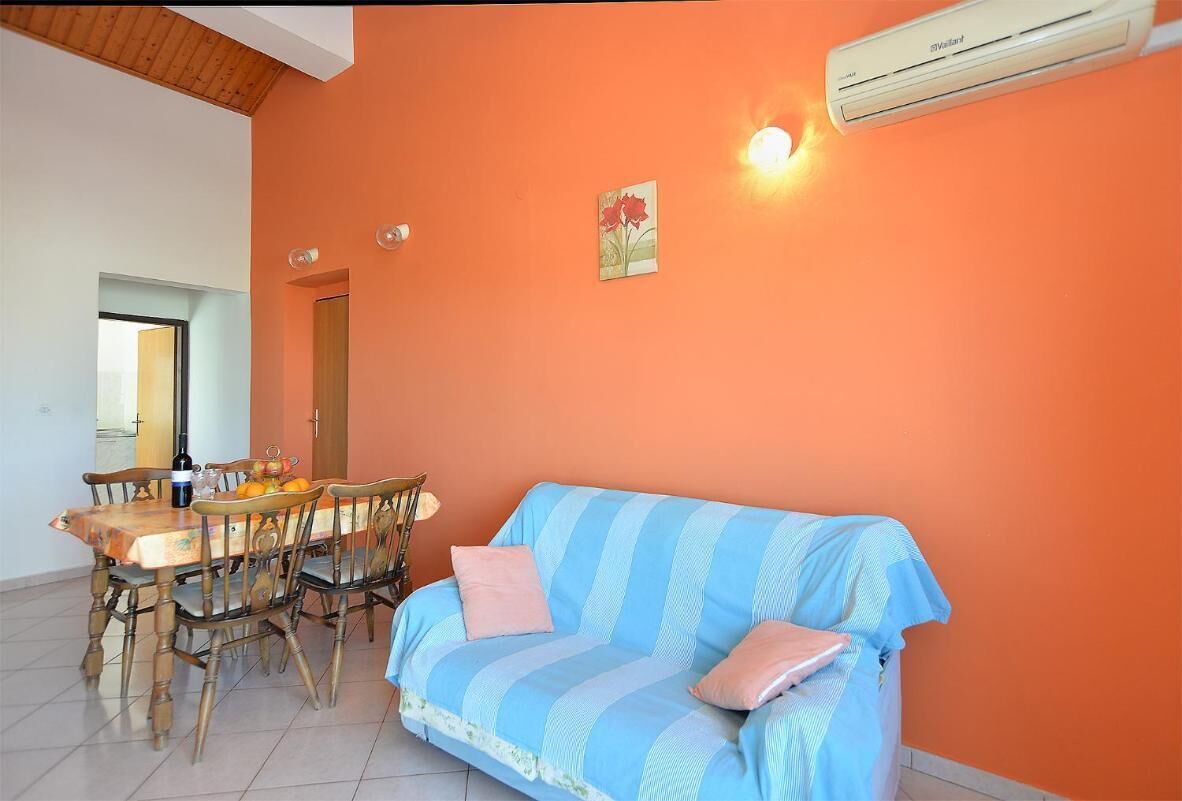 A dining area with a wooden table and chairs, a blue-striped sofa, and an orange wall with a painting and an air conditioning unit.
