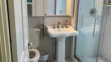 Une salle de bain avec un lavabo sur colonne blanc, des toilettes, une douche en verre et un sol carrelé.