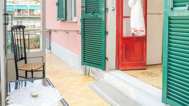 Un balcón con una silla de mimbre, una mesita con un cuenco y una puerta roja con contraventanas verdes.