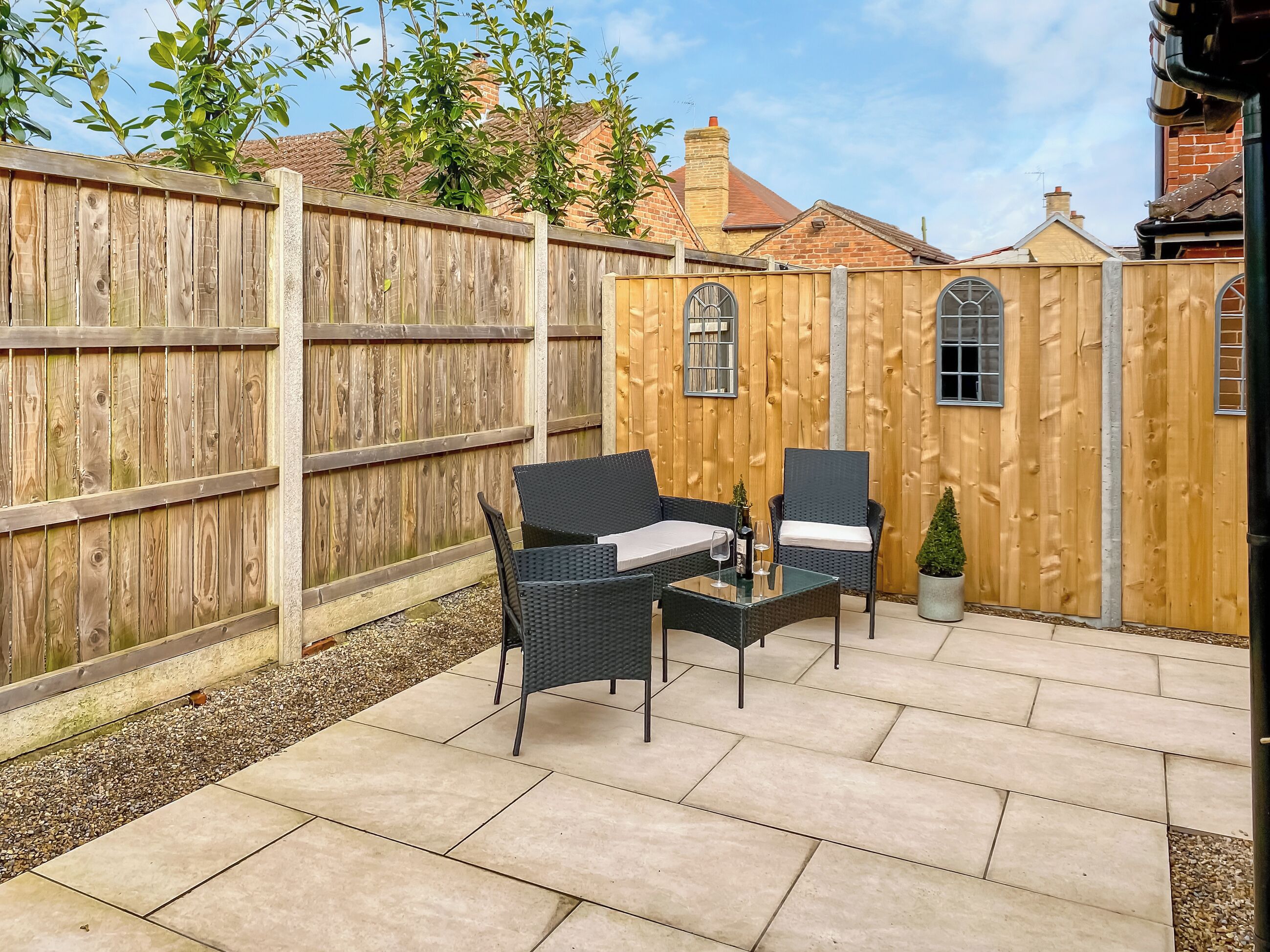 A patio with two wicker chairs and a table, a wooden fence, and a brick building in the background.