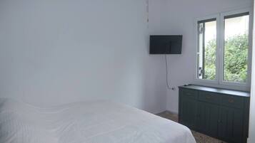 A bedroom with a bed, a dresser, a TV mounted on the wall, and a window with a view of greenery outside.
