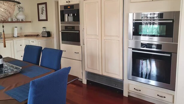 A kitchen with white cabinetry, a built-in oven, and a microwave. There is a dining area with blue chairs and a table set for two.