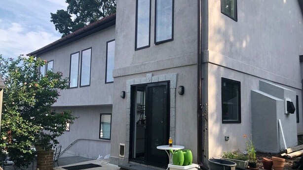 Ein modernes, zweistöckiges Haus mit einer großen Haustür, einer kleinen Terrasse mit Tisch und Stühlen und einem Gartenbereich mit Blumentöpfen.