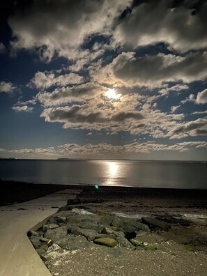 A coastal scene with a rocky foreground, a calm sea, and a sky with clouds and the sun.