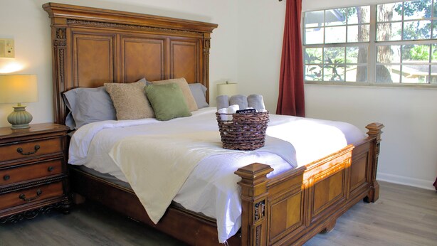 A wooden bed with a white comforter and pillows, a bedside table with a lamp, and a window with a view of trees. - SHADOW WOOD - King Size Private Suite Sleeps 4, Near Universal, I-Drive, Disney (Pine Hills)