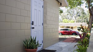 A beige brick house with a white door, a red car parked in the driveway, and potted plants on the porch. - SHADOW WOOD - King Size Private Suite Sleeps 4, Near Universal, I-Drive, Disney (Pine Hills)