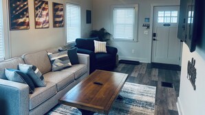 A living room with a gray sofa, a wooden coffee table, and a TV mounted on the wall. There are three framed American flags on the wall, and a door with a 'No Smoking' sign.