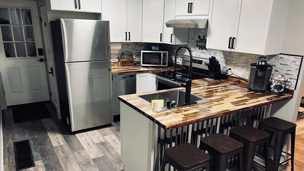 A modern kitchen with white cabinets, a stainless steel refrigerator, a microwave, a sink, and wooden bar stools.
