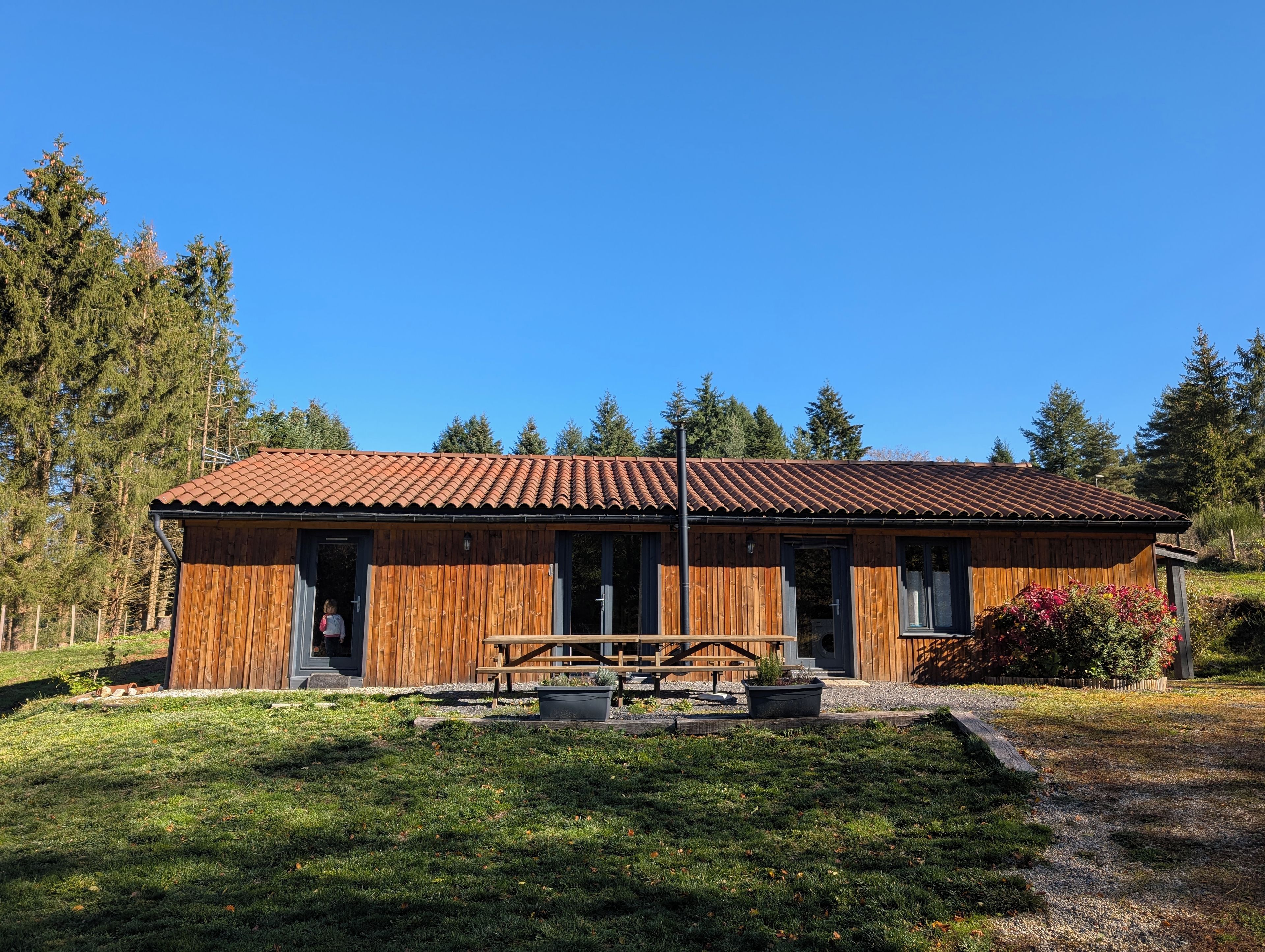 A wooden cabin with a tiled roof, a picnic table, and a small garden area.