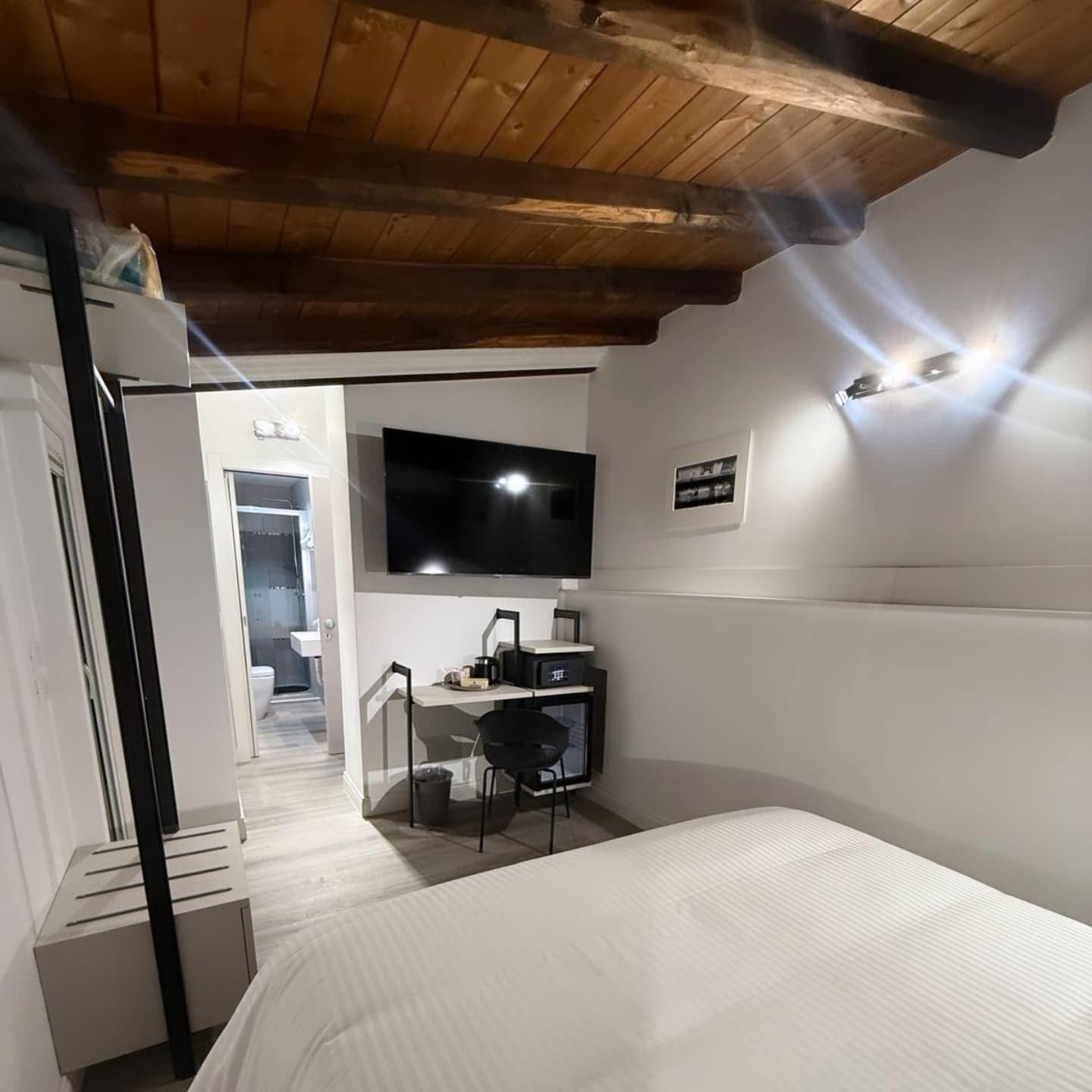 A compact hotel room with a wooden ceiling, a bed, a desk with a chair, a television mounted on the wall, and a small window.