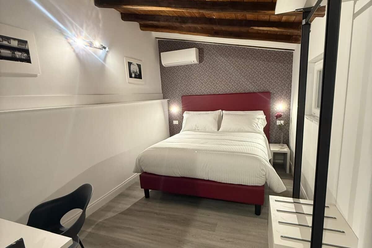 A bedroom with a wooden ceiling, a bed with white bedding, a desk with a chair, and a bedside table.