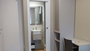 A room with a wooden floor, a bed with a green cover, a desk with a chair, a wardrobe, and a bathroom with a sink and mirror. - Vroom509 (Verona)