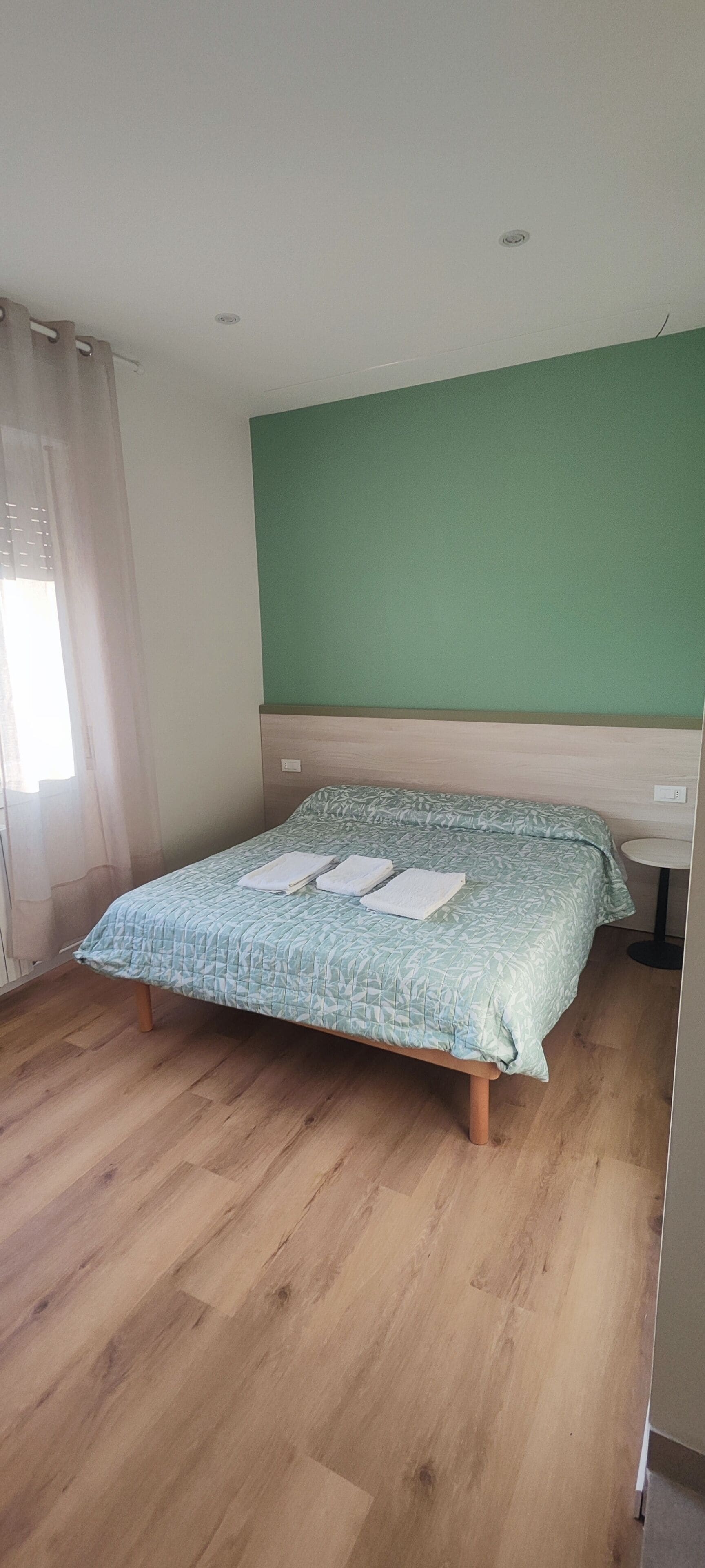 A single bed with a patterned bedspread, a round table, and a green accent wall in a room with wooden flooring.