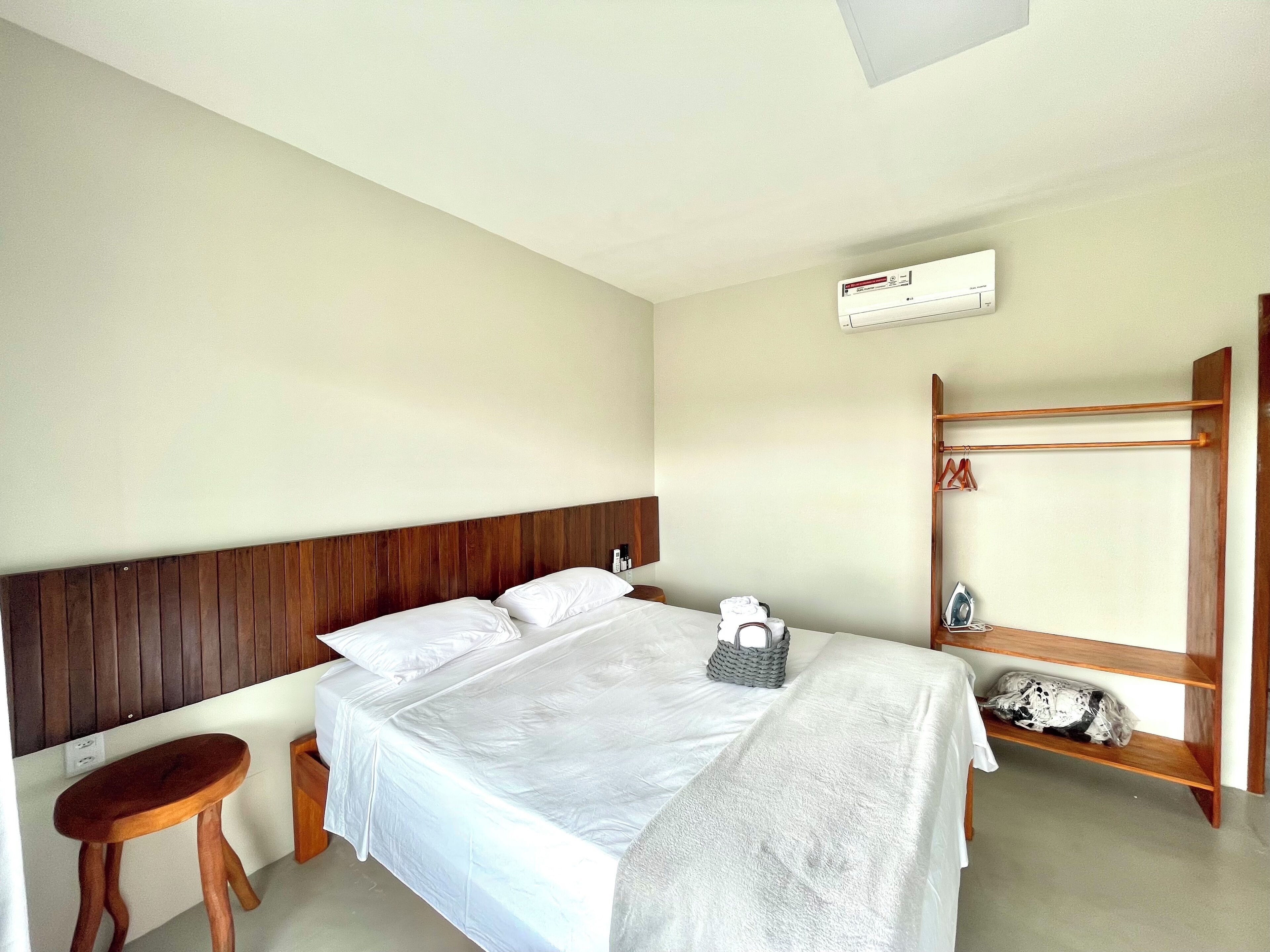 A hotel room with a bed, a wooden headboard, a small wooden stool, a mirror, and a shelf with a towel and an iron.