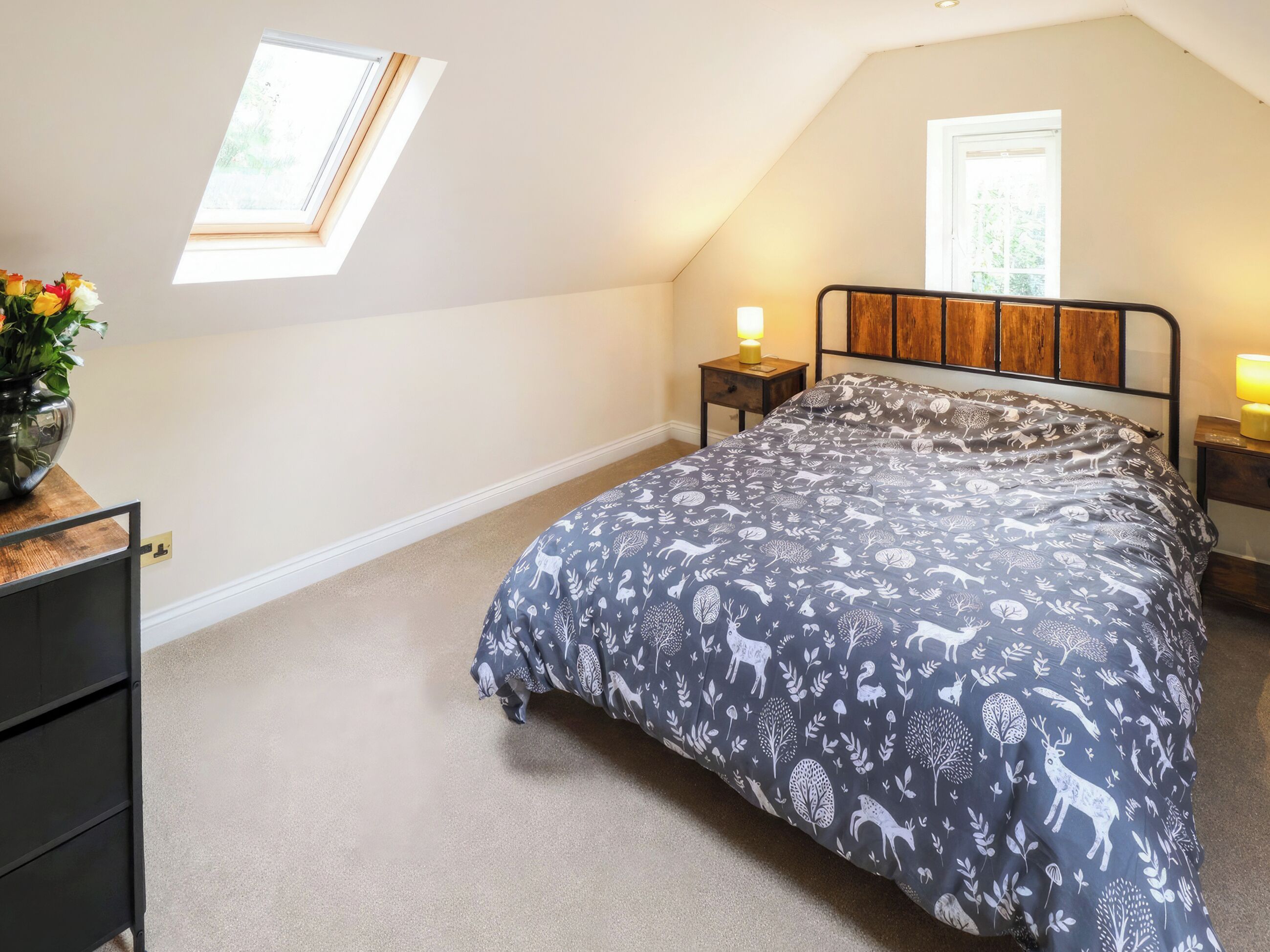 A bedroom with a sloped ceiling, a skylight, a bed with a patterned duvet, bedside tables with lamps, and a small wooden nightstand.