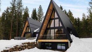 Uma cabana moderna em formato triangular, com grandes janelas e base de pedra, cercada por neve e árvores.