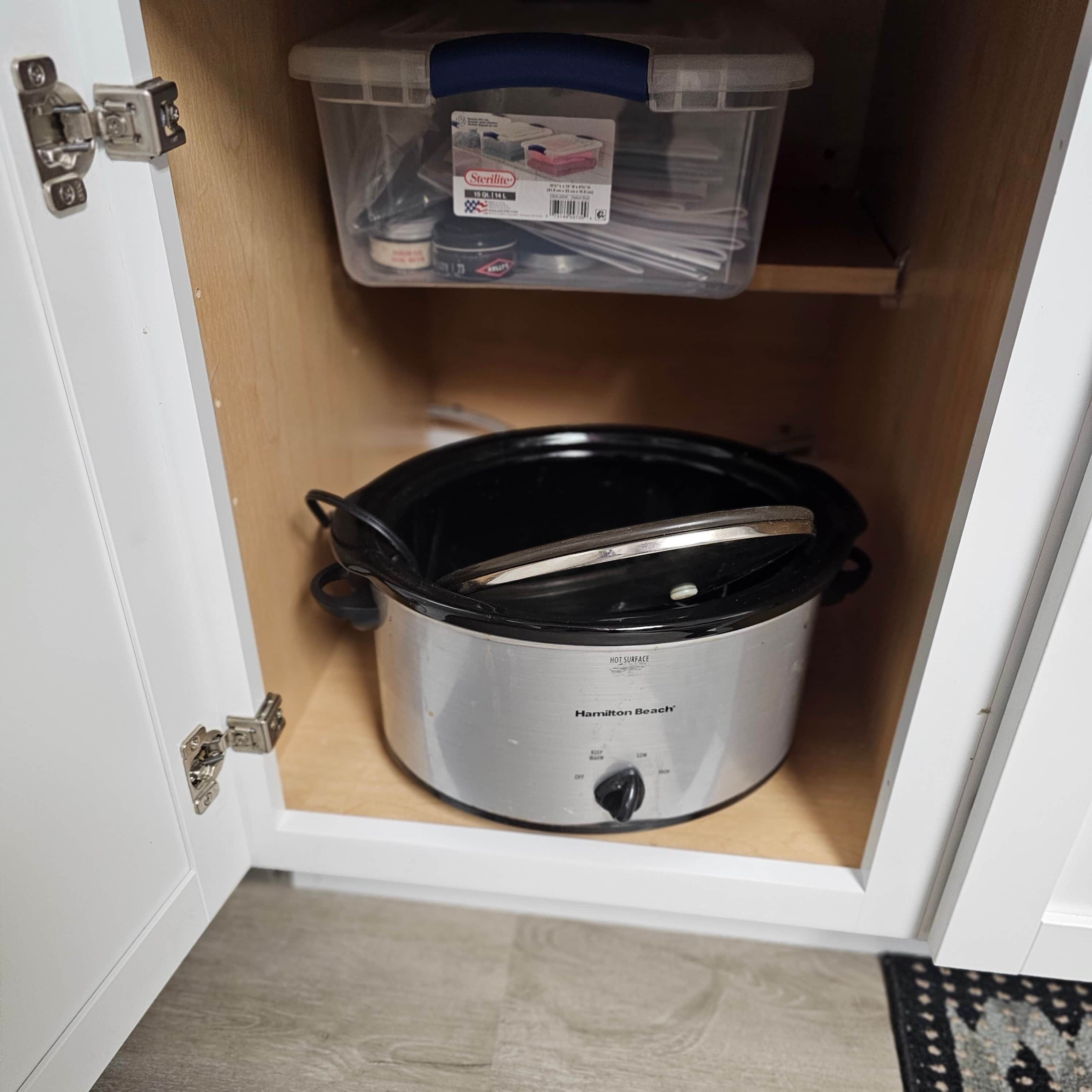 Una cocina con una olla de cocción lenta, un recipiente de plástico con objetos y un estante de madera.