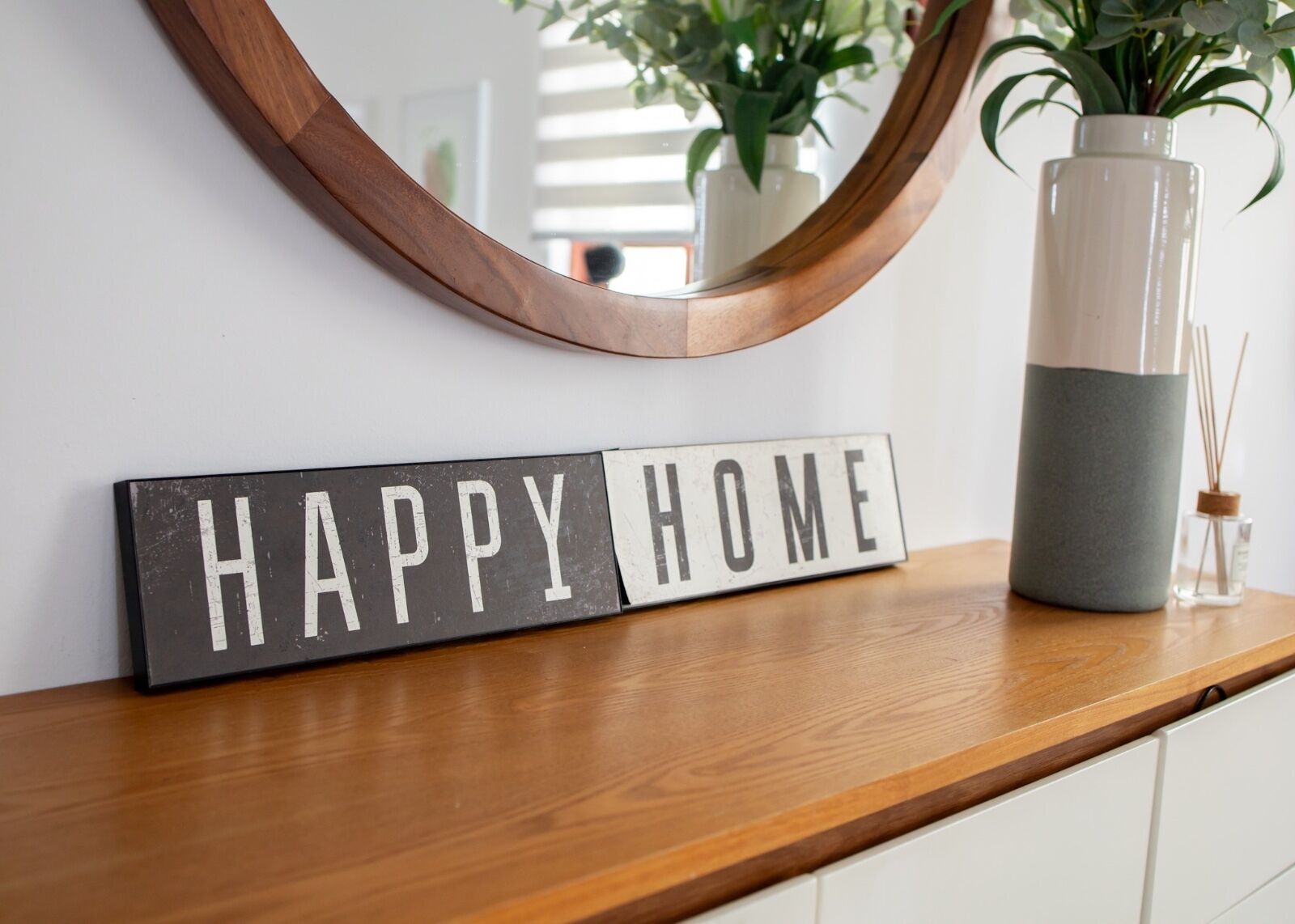 A sign with the words "HAPPY HOME" on a wooden surface, a mirror, and a vase with plants.