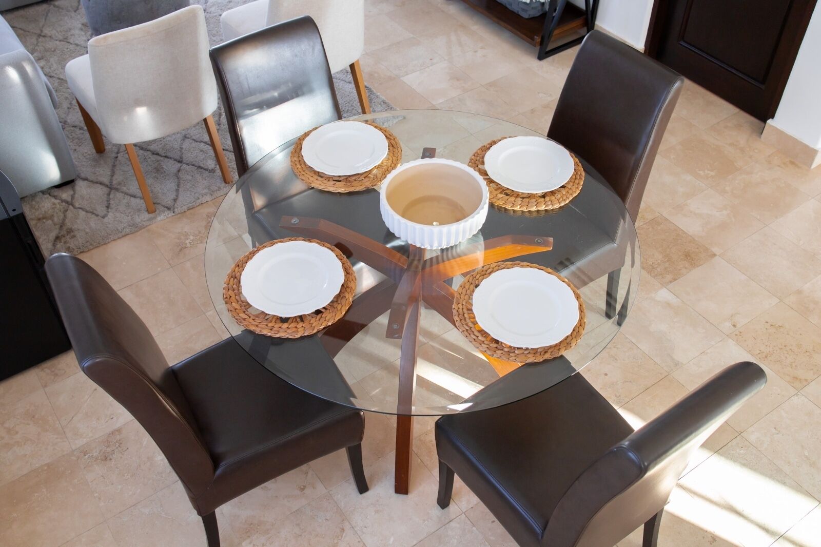 A round glass table with woven placemats and white plates, surrounded by dark leather chairs in a well-lit room with tiled flooring.