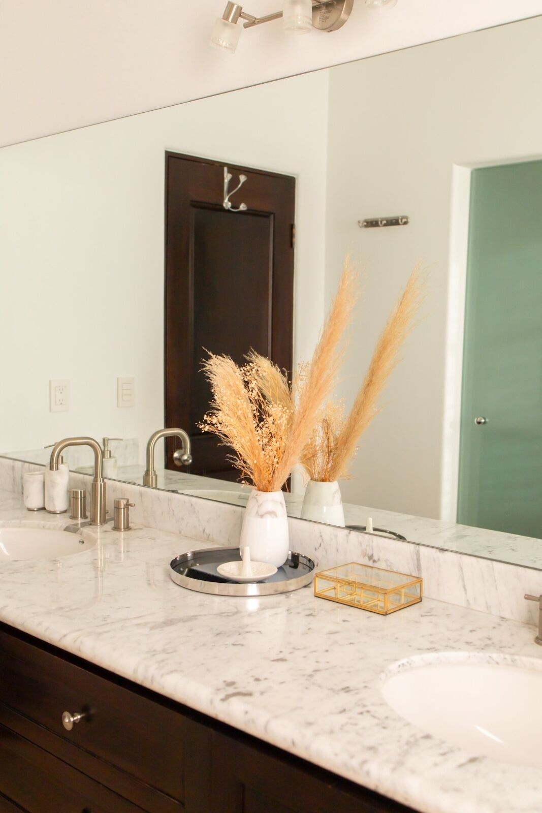A bathroom with a marble countertop, a sink, a mirror, a door, and a vase with pampas grass.