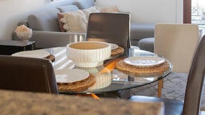 Een moderne woonkamer met een glazen salontafel, een grijze bank en een eettafel met borden en kommen.