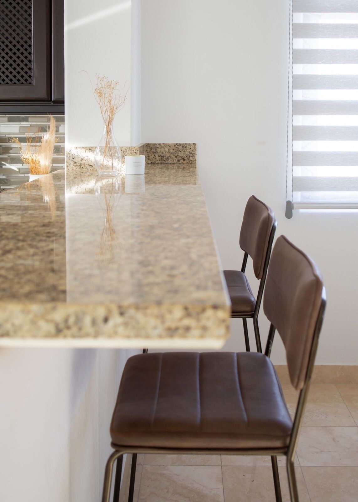 A modern kitchen with a granite countertop, a white vase with dried flowers, and a window with blinds.