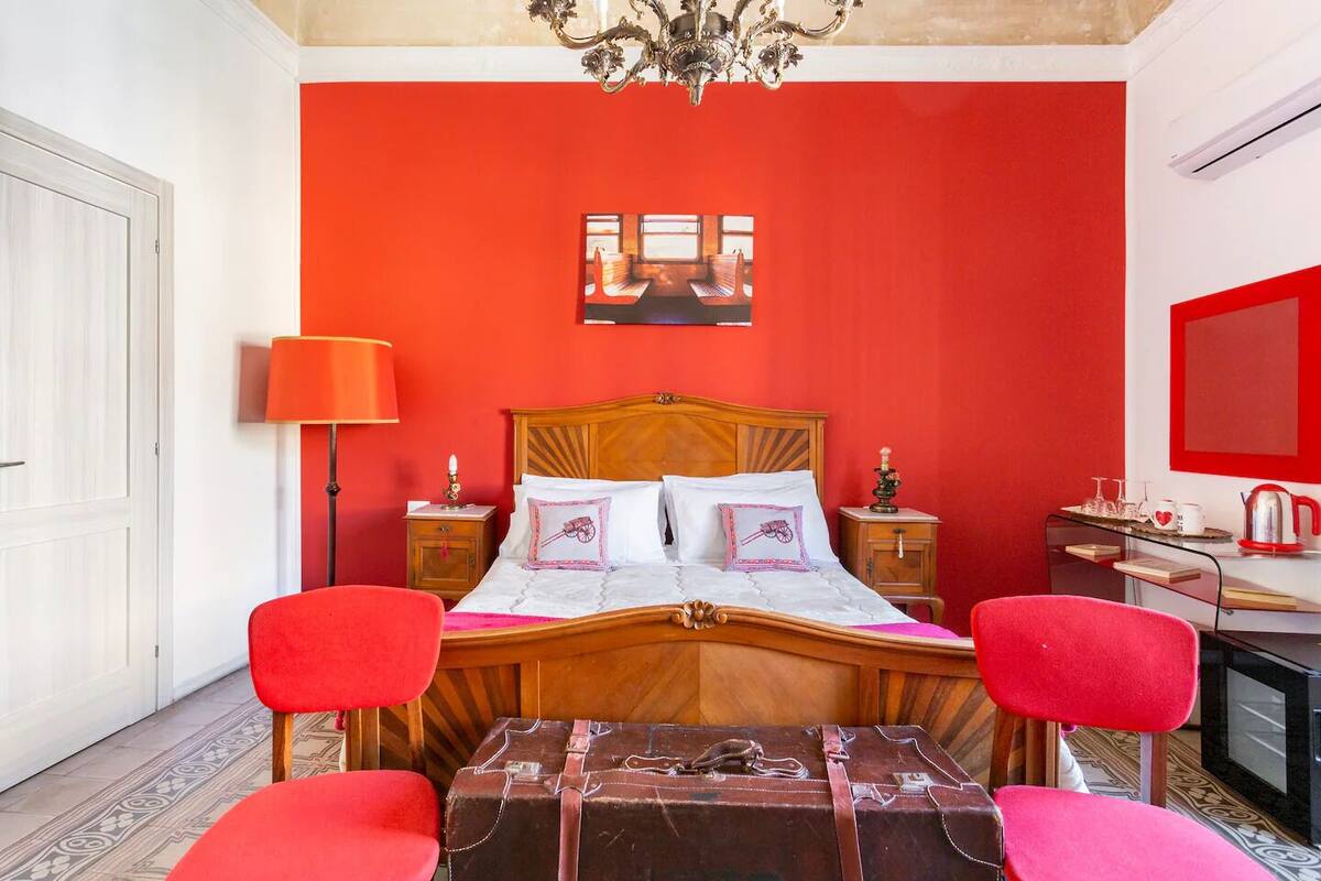A bedroom with a red wall, a wooden bed, two bedside tables, a desk with a chair, and a suitcase on the floor.