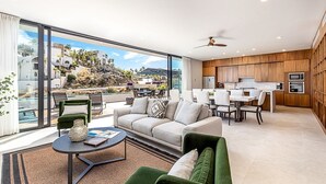 A modern living room with a large sliding glass door leading to a balcony, a sofa, a coffee table, and a dining area with a kitchen in the background.