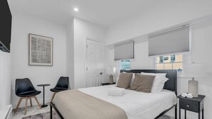 A modern hotel room with a bed, two chairs, a nightstand, and a window with blinds. - Budget-Friendly Apt | WD, Wifi & Modern Amenities (Paterson)