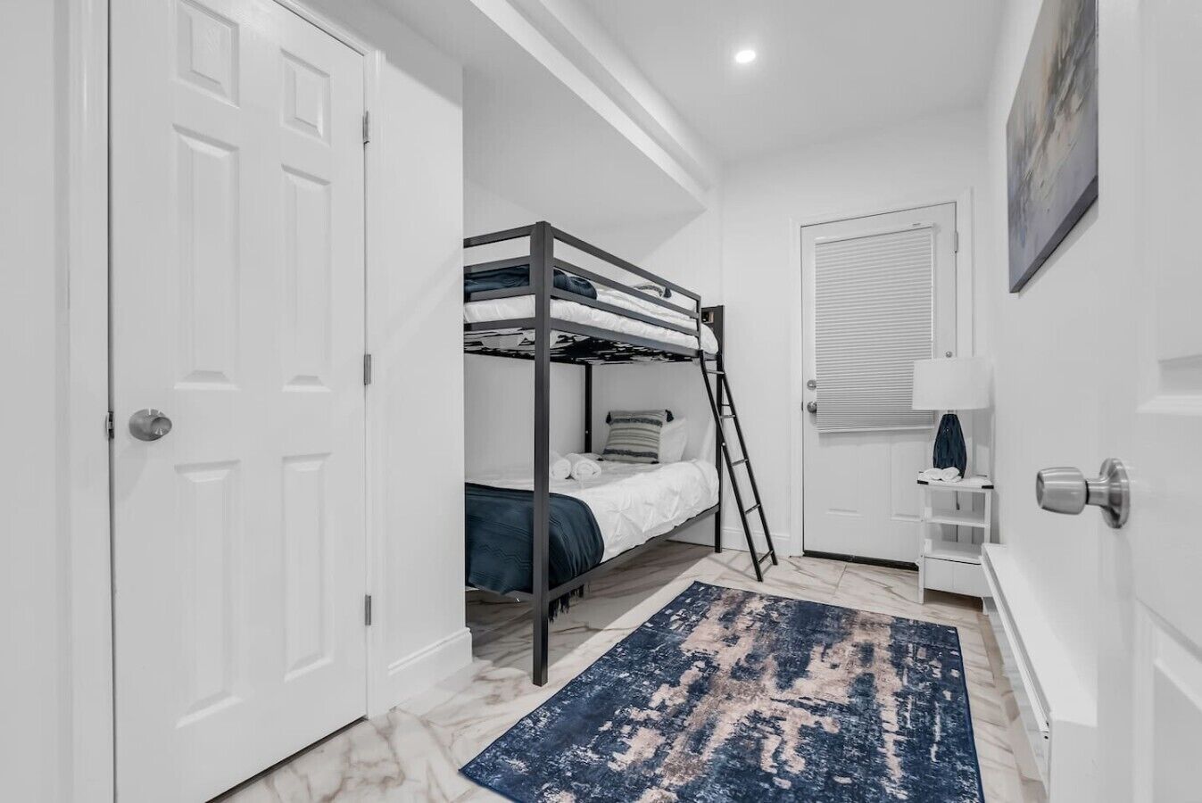 A bedroom with a bunk bed, a ladder, a door with a handle, a white wall, and a blue rug.