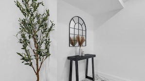 A room with a tall potted plant, a black console table with a vase, and a window with a geometric design. - Budget-Friendly Apt | WD, Wifi & Modern Amenities (Paterson)