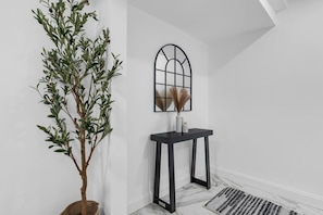 Een kamer met een hoge kamerplant, een zwarte consoles en een raam met een geometrisch ontwerp.