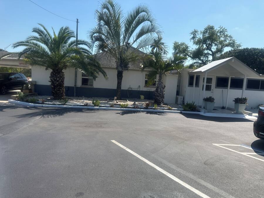 A parking lot with a building that has a white exterior and a sloped roof. There are palm trees in front of the building.