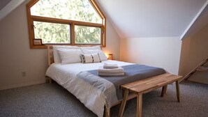 A bedroom with a large window, a bed with a gray blanket, a wooden bench, and a small bedside table.