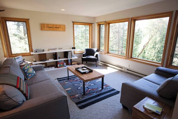 Een moderne woonkamer met een grijze bank, een zwarte fauteuil en een houten salontafel. Er zijn grote ramen met uitzicht op bomen.