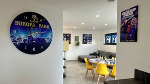 A modern hotel room with a large clock featuring a cityscape, a dining area with yellow chairs, and a sofa. - Europ-Appart ~ {Europa-Park / Rulantica} (Huttenheim)