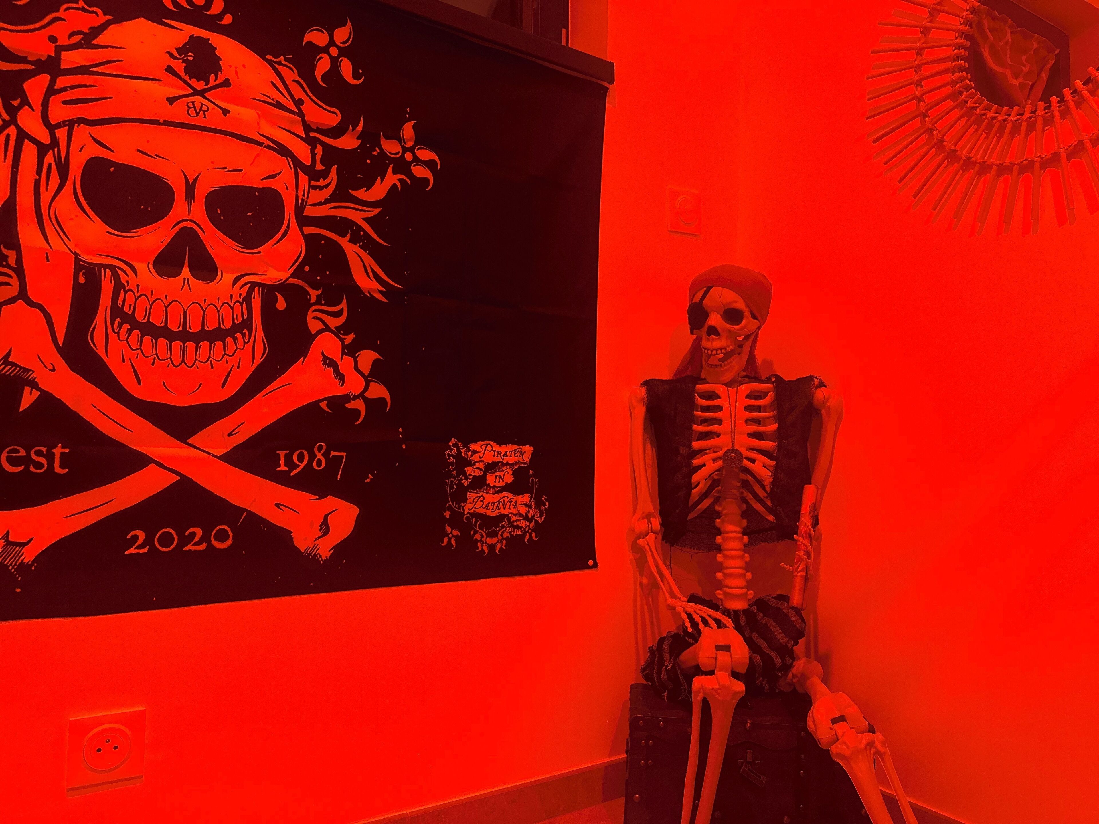 Un esqueleto vestido con chaleco y sombrero, sentado sobre una maleta frente a una pared de fondo rojo y con un gráfico de calavera.