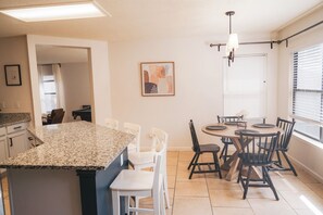 Eine moderne Küche mit Granitabdeckung, weißen Stühlen, einem Esstisch mit schwarzen Stühlen, einer Pendelleuchte und einem gerahmten Kunstwerk an der Wand.