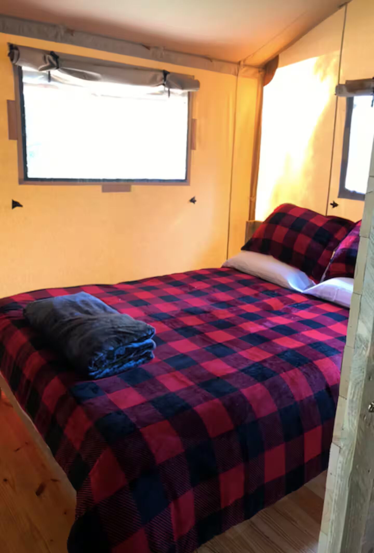A single bed with a red and black checkered bedspread, a folded blanket, and a pillow. There is a window above the bed with curtains, and a wooden door to the right.