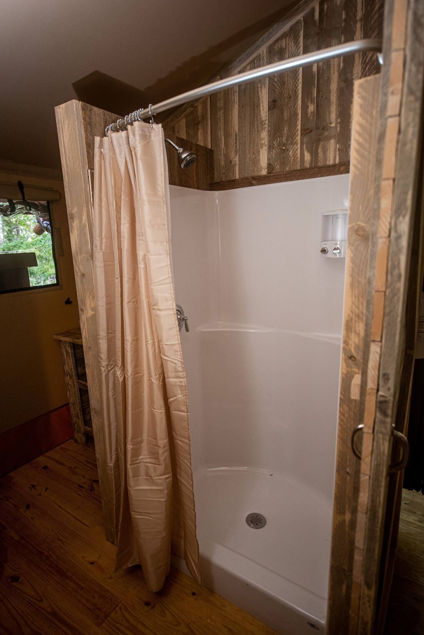 A shower area with a wooden wall, a beige shower curtain, and a window with a view of greenery.