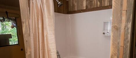 A shower area with a wooden wall, a beige shower curtain, and a window with a view of greenery.