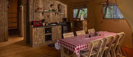 Un intérieur chaleureux et rustique, avec une table à manger en bois disposée pour quatre personnes, un coin cuisine avec un micro-ondes rouge et une fenêtre donnant sur un espace verdoyant.