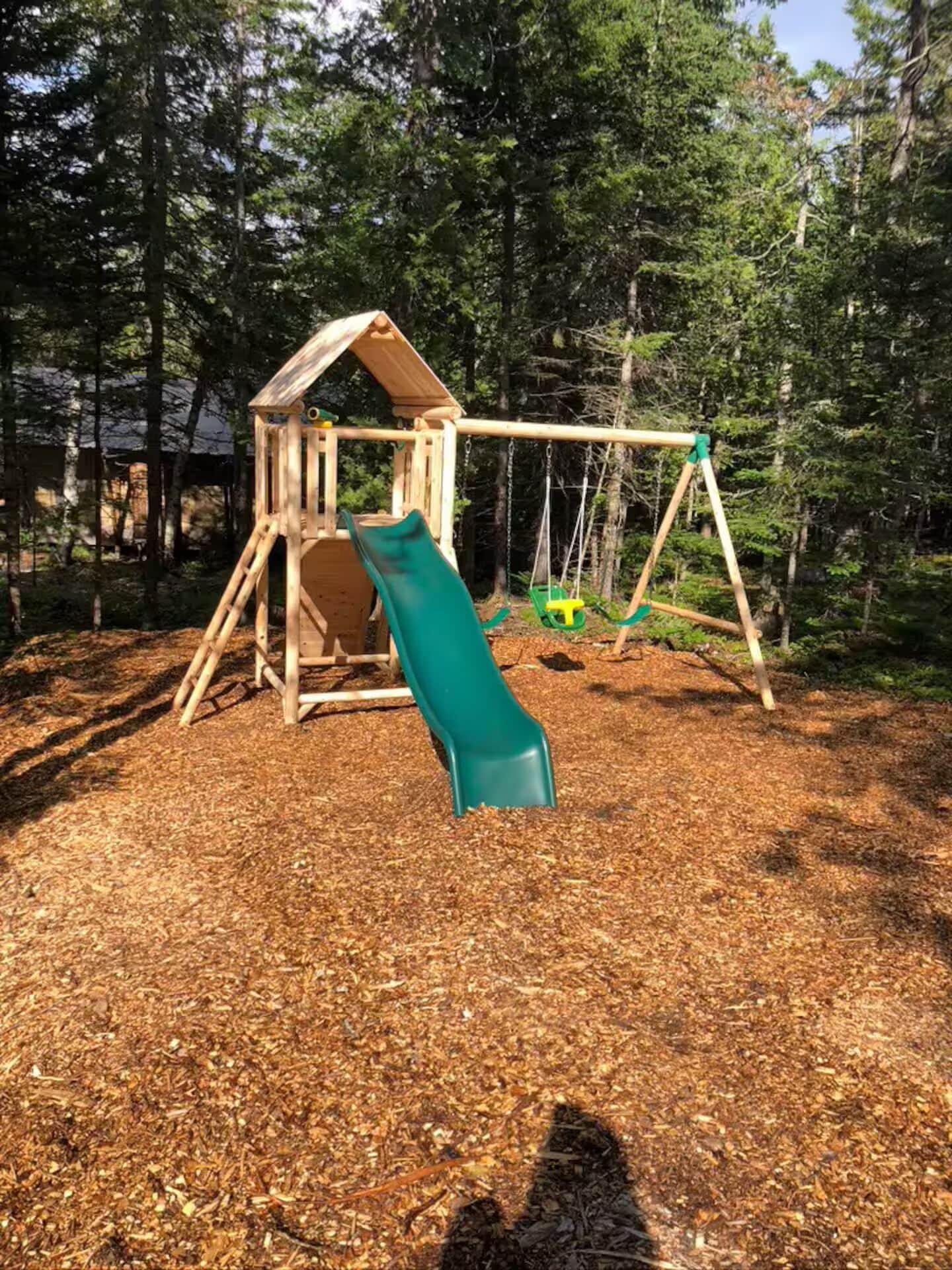 A wooden play structure with a slide and swings in a wooded area.